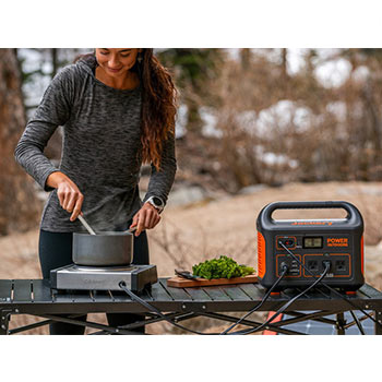 A jackery explorer 1000 solar generator beside a woman using an electric cooker outdoors